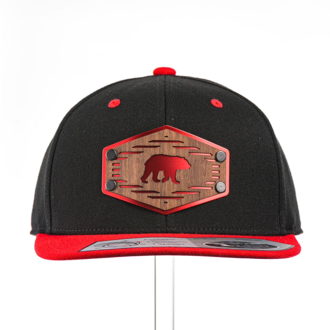 The front of a Grizzly Bear w/ Lines Walnut Wood Black & Red Flat Bill Hat, featuring a hexagonal real walnut wood veneer patch with a cut-out bear silhouette and fine line details, laminated to a hexagonal red anodized aluminum plate, and riveted to a black and red Flexfit 110F flat bill hat—handmade in Montana, USA by Last Best Supply Co.
