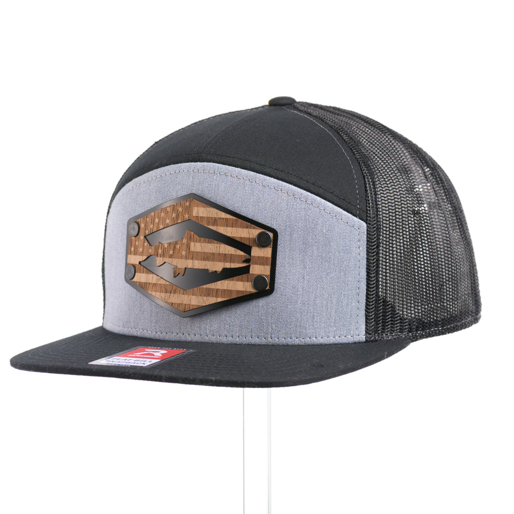 3/4 view of a Walnut Wood Chinook Salmon American Flag Flat Bill Trucker Cap, featuring a real walnut wood veneer patch precision-cut into a hexagon, etched with an American flag with a Chinook salmon cut-out, laminated to a hexagonal black anodized aluminum plate, and riveted to a heather gray and black Richardson 168 flat bill trucker hat—handmade in Montana, USA by Last Best Supply Co.