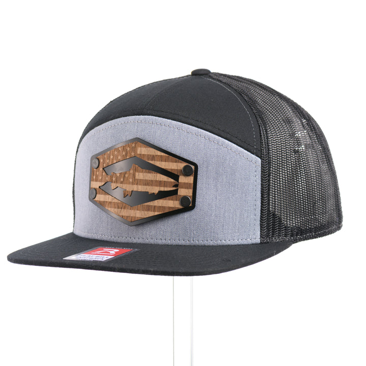 3/4 view of a Walnut Wood Chinook Salmon American Flag Flat Bill Trucker Cap, featuring a real walnut wood veneer patch precision-cut into a hexagon, etched with an American flag with a Chinook salmon cut-out, laminated to a hexagonal black anodized aluminum plate, and riveted to a heather gray and black Richardson 168 flat bill trucker hat—handmade in Montana, USA by Last Best Supply Co.