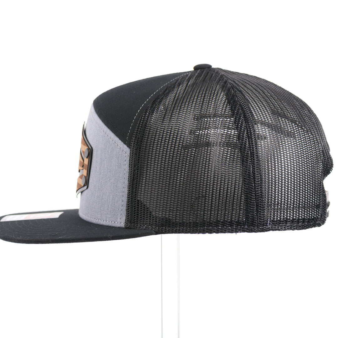 The side of a Walnut Wood Chinook Salmon American Flag Flat Bill Trucker Cap, featuring a real walnut wood veneer patch precision-cut into a hexagon, etched with an American flag with a Chinook salmon cut-out, laminated to a hexagonal black anodized aluminum plate, and riveted to a heather gray and black Richardson 168 flat bill trucker hat—handmade in Montana, USA by Last Best Supply Co.