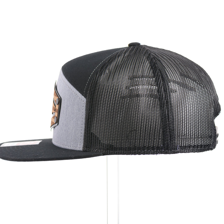 The side of a Walnut Wood Chinook Salmon American Flag Flat Bill Trucker Cap, featuring a real walnut wood veneer patch precision-cut into a hexagon, etched with an American flag with a Chinook salmon cut-out, laminated to a hexagonal black anodized aluminum plate, and riveted to a heather gray and black Richardson 168 flat bill trucker hat—handmade in Montana, USA by Last Best Supply Co.