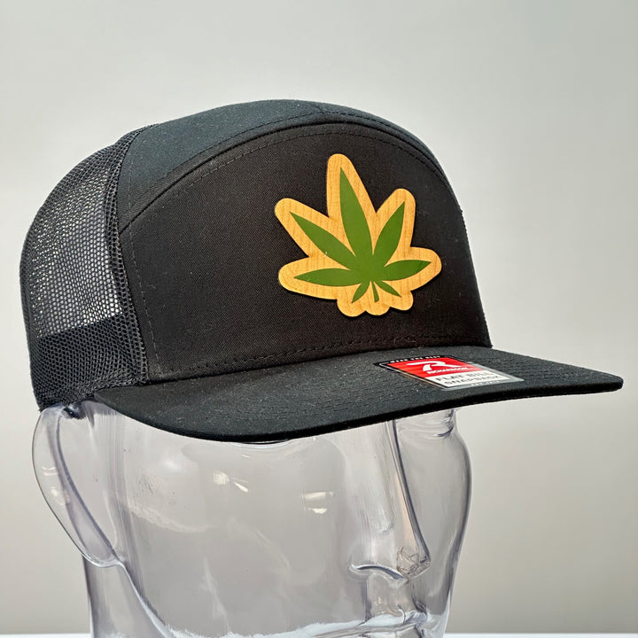 3/4 view of a Hemp Leaf Flexwood Patch Black Flat Bill Trucker Hat, featuring a Flexwood veneer patch cut into the shape of a hemp leaf, and printed with green ink, securely attached to a solid black Richardson 112 flat bill trucker hat—handmade in Montana, USA by Last Best Supply Co.