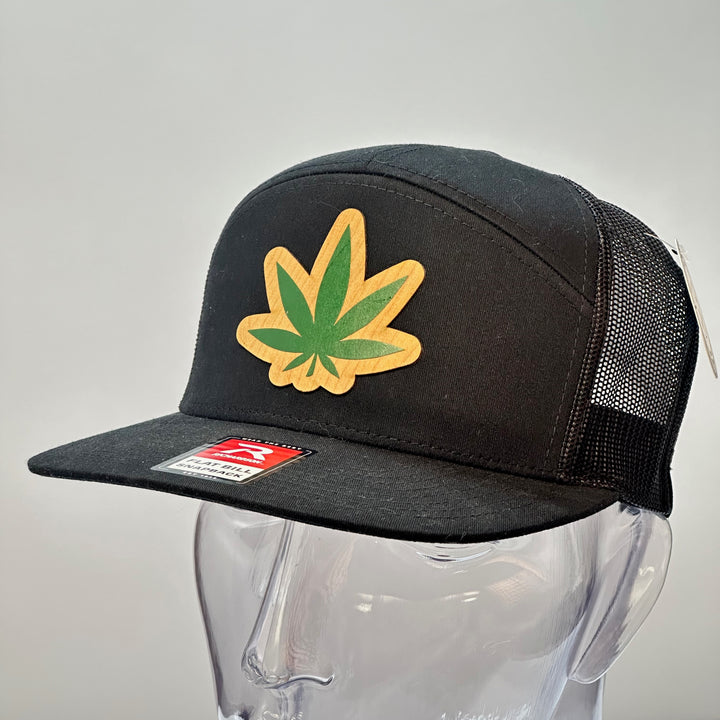 Hemp Leaf Flexwood Patch Black Flat Bill Trucker Hat, featuring a Flexwood veneer patch cut into the shape of a hemp leaf, and printed with green ink, securely attached to a solid black Richardson 112 flat bill trucker hat—handmade in Montana, USA by Last Best Supply Co.