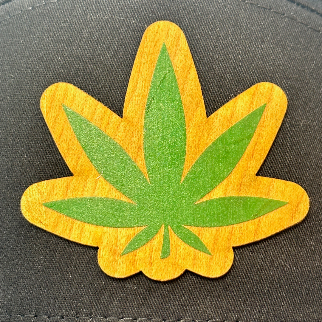 Patch detail of a Hemp Leaf Flexwood Patch Black Flat Bill Trucker Hat, featuring a Flexwood veneer patch cut into the shape of a hemp leaf, and printed with green ink, securely attached to a solid black Richardson 112 flat bill trucker hat—handmade in Montana, USA by Last Best Supply Co.