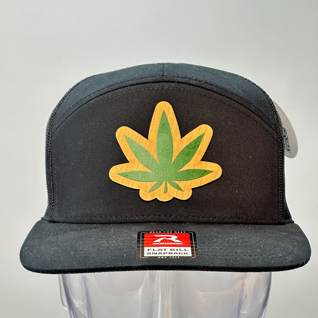 Front of a Hemp Leaf Flexwood Patch Black Flat Bill Trucker Hat, featuring a Flexwood veneer patch cut into the shape of a hemp leaf, and printed with green ink, securely attached to a solid black Richardson 112 flat bill  trucker hat—handmade in Montana, USA by Last Best Supply Co.