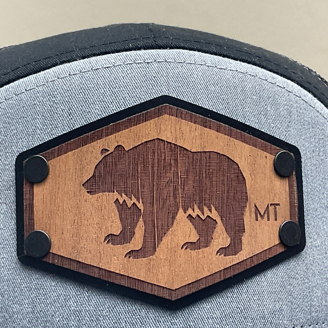 Patch detail of a Hex Bear MT Wood Patch Richardson Flat Bill Hat, featuring a hexagonal, real wood veneer patch etched with a grizzly bear and the letters “MT”, laminated to a black anodized metal plate, and then riveted to heather gray and black Richardson 168 seven-panel flat bill trucker hat—handmade in Montana, USA by Last Best Supply Co.