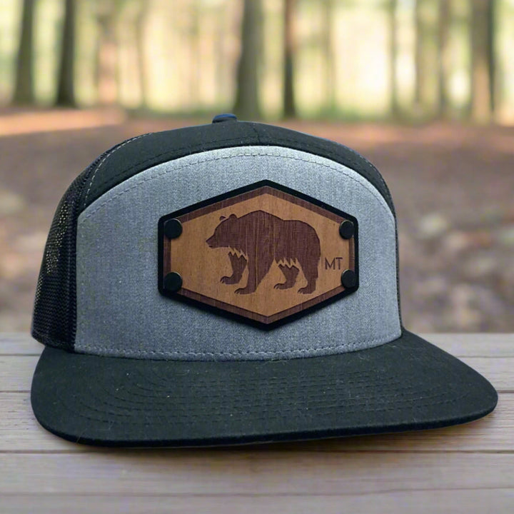 Front of a Hex Bear MT Wood Patch Richardson Flat Bill Hat, featuring a hexagonal, real wood veneer patch etched with a grizzly bear and the letters “MT”, laminated to a black anodized metal plate, and then riveted to heather gray and black Richardson 168 seven-panel flat bill trucker hat—handmade in Montana, USA by Last Best Supply Co.