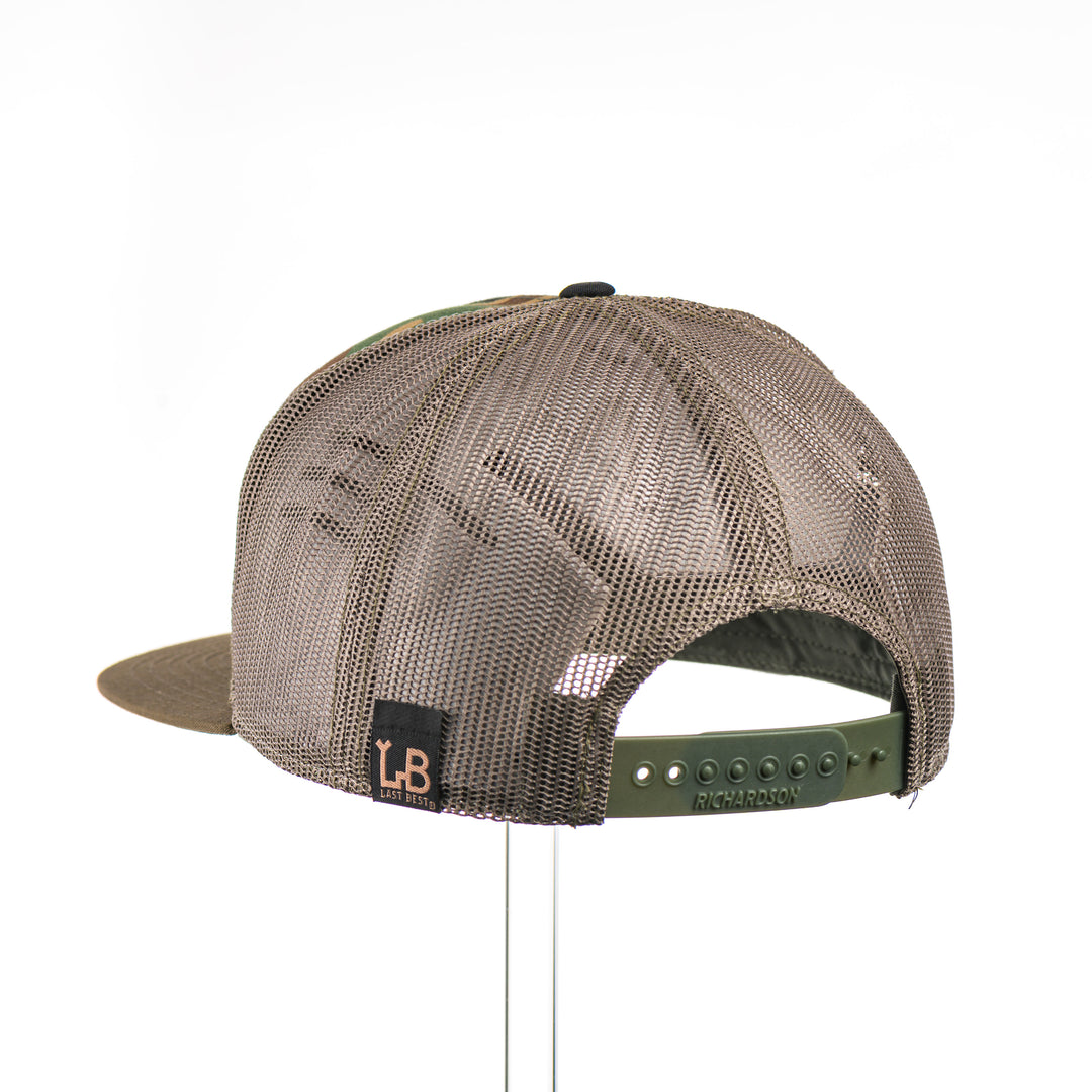 The back of a Mahogany Wood Bigfoot American Flag Patch Camo Flat Bill Hat, featuring a hexagonal real mahogany wood veneer patch etched with an American flag with cut-out Bigfoot silhouette, laminated to a hexagonal black anodized aluminum plate, and riveted to a black, camo, and loden green Richardson 168 seven-panel trucker hat—handmade in Montana, USA by Last Best Supply Co.