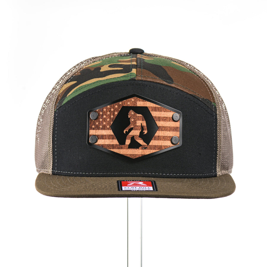 The front of a Mahogany Wood Bigfoot American Flag Patch Camo Flat Bill Hat, featuring a hexagonal real mahogany wood veneer patch etched with an American flag with cut-out Bigfoot silhouette, laminated to a hexagonal black anodized aluminum plate, and riveted to a black, camo, and loden green Richardson 168 seven-panel trucker hat—handmade in Montana, USA by Last Best Supply Co.