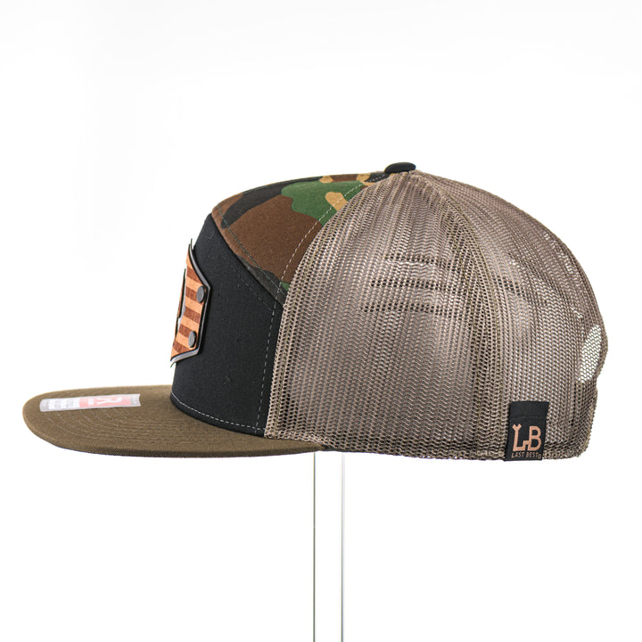 The side of a Mahogany Wood Bigfoot American Flag Patch Camo Flat Bill Hat, featuring a hexagonal real mahogany wood veneer patch etched with an American flag with cut-out Bigfoot silhouette, laminated to a hexagonal black anodized aluminum plate, and riveted to a black, camo, and loden green Richardson 168 seven-panel trucker hat—handmade in Montana, USA by Last Best Supply Co.