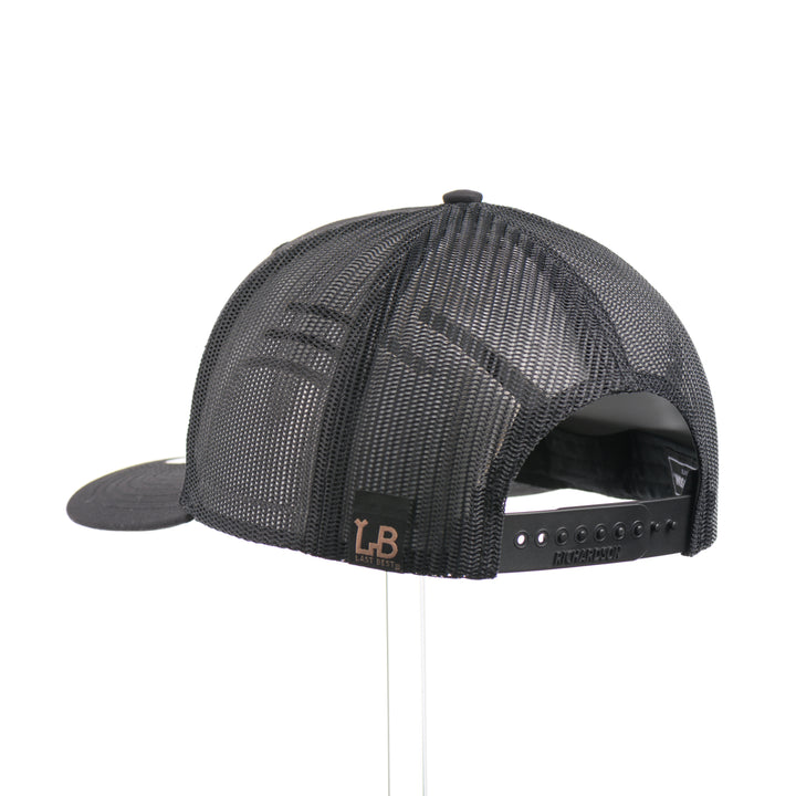 The back of a black Richardson trucker hat with an etched mahogany wood American flag and bear cut out on a black metal patch, handmade in Montana USA by Last Best Supply Co.