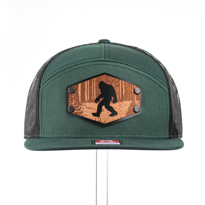The front of a Mahogany Wood Roaming Bigfoot Patch Flat Bill Trucker Hat, featuring a real mahogany wood veneer patch precision-cut into a hexagon, etched with a forest scene with a silhouette of a roaming Sasquatch, laminated to a hexagon black anodized aluminum plate, and riveted to a dark forest green and black mesh Richardson 168 flat bill trucker hat—handmade in Montana, USA by Last Best Supply Co.