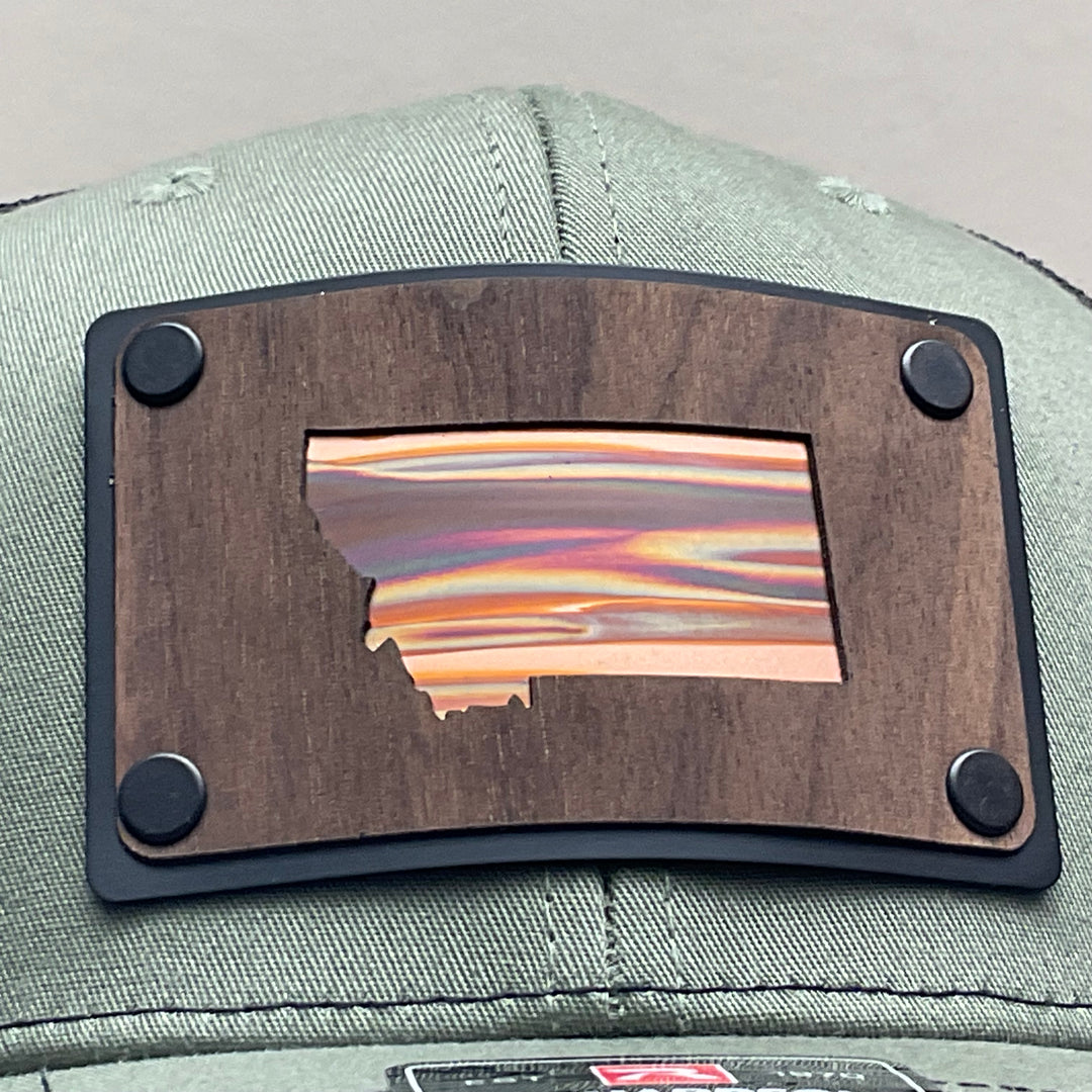 Patch detail of a Montana Walnut Wood with Sundae Copper Patch on Richardson Trucker Hat, featuring a rectangular, real walnut wood veneer patch with a precision-cut state of Montana silhouette cutout, laminated to a black anodized metal plate, and then riveted to an olive green Richardson 112 trucker hat with black mesh—handmade in Montana, USA by Last Best Supply Co.