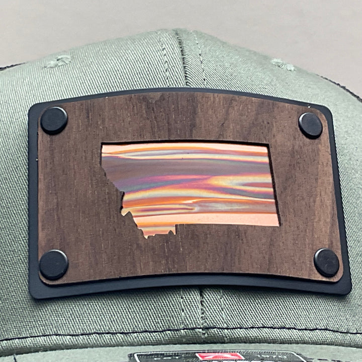 Patch detail of a Montana Walnut Wood with Sundae Copper Patch on Richardson Trucker Hat, featuring a rectangular, real walnut wood veneer patch with a precision-cut state of Montana silhouette cutout, laminated to a black anodized metal plate, and then riveted to an olive green Richardson 112 trucker hat with black mesh—handmade in Montana, USA by Last Best Supply Co.