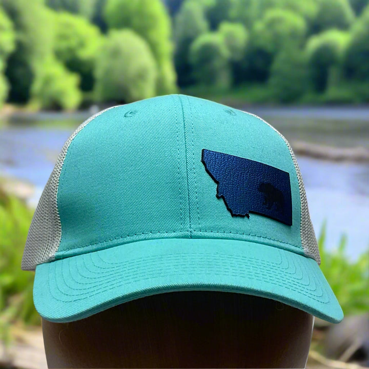 Front of a Premium Leather Montana Bear on Low Profile Teal Trucker Hat, featuring a blue hand-dyed leather patch cut in the shape of the state of Montana with a grizzly bear cut-out, heat pressed to the structured front of a teal and white  trucker hat—handmade in Montana, USA by Last Best Supply Co.