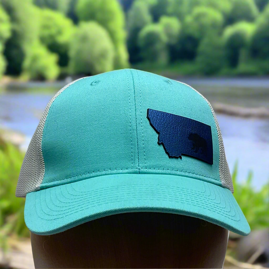 Front of a Premium Leather Montana Bear on Low Profile Teal Trucker Hat, featuring a blue hand-dyed leather patch cut in the shape of the state of Montana with a grizzly bear cut-out, heat pressed to the structured front of a teal and white  trucker hat—handmade in Montana, USA by Last Best Supply Co.