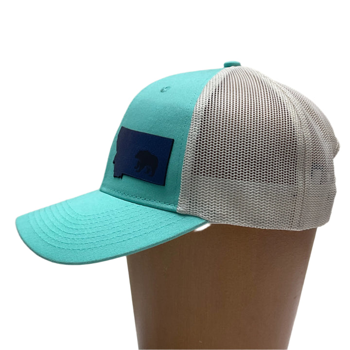Side view of a Premium Leather Montana Bear on Low Profile Teal Trucker Hat, featuring a blue hand-dyed leather patch cut in the shape of the state of Montana with a grizzly bear cut-out, heat pressed to the structured front of a teal and white  trucker hat—handmade in Montana, USA by Last Best Supply Co.