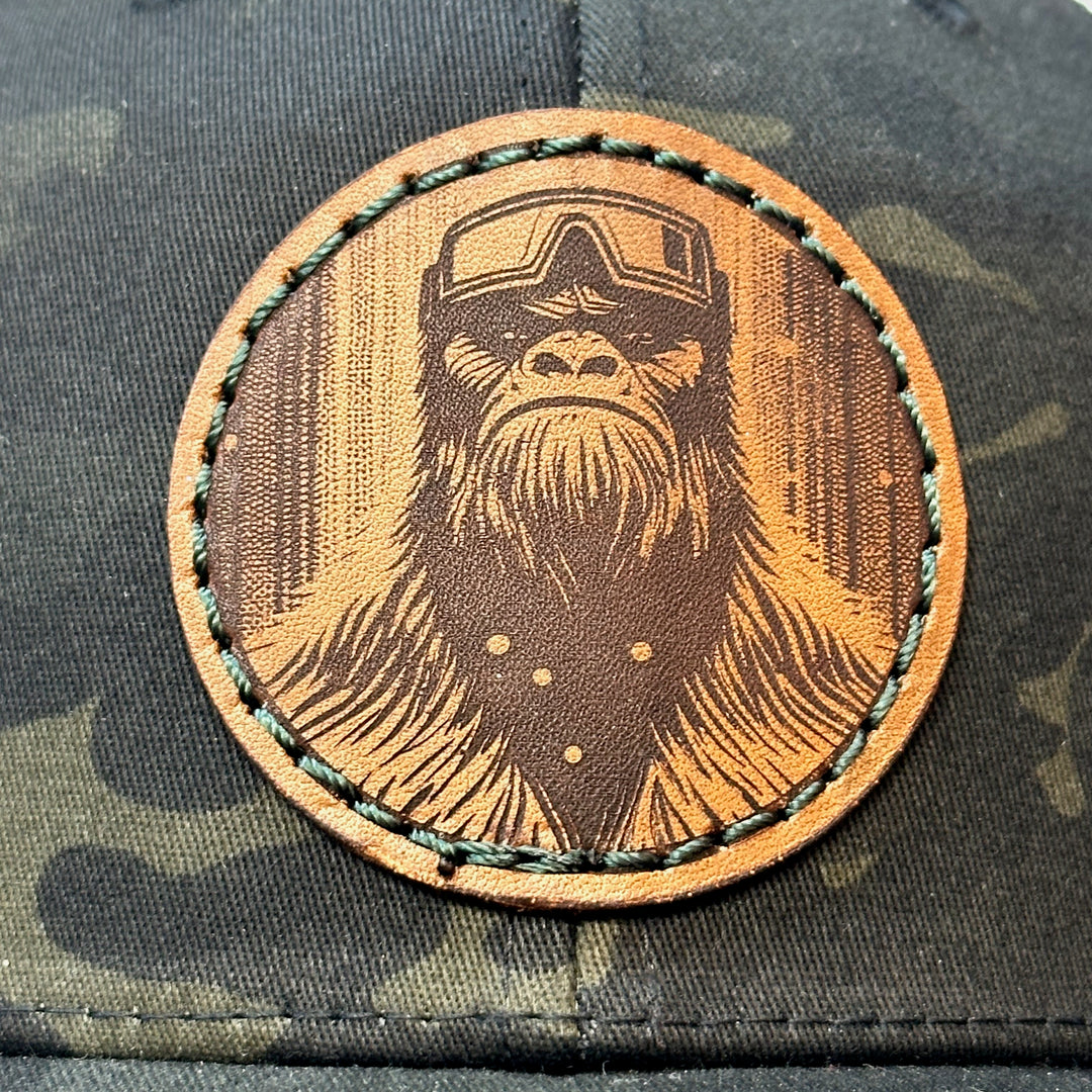 Detail of Snowy Bigfoot Leather Patch Black Camo Trucker Hat, featuring a circular real hand-dyed leather patch etched with a detailed Bigfoot in ski googles design, securely heat bonded and sewn onto the structured front of a dark camo Yupoong Retro Trucker Hat—handmade in Montana, USA by Last Best Supply Co.