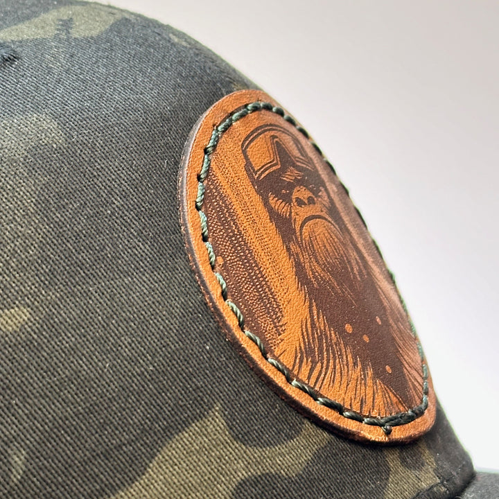 Detail of Snowy Bigfoot Leather Patch Black Camo Trucker Hat, featuring a circular real hand-dyed leather patch etched with a detailed Bigfoot in ski googles design, securely heat bonded and sewn onto the structured front of a dark camo Yupoong Retro Trucker Hat—handmade in Montana, USA by Last Best Supply Co.