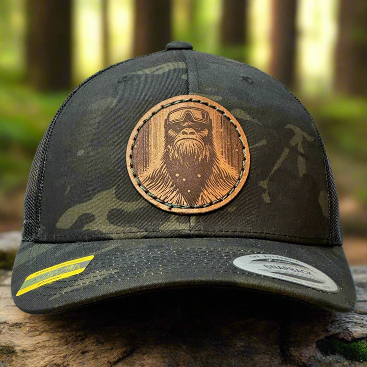 The front of a Snowy Bigfoot Leather Patch Black Camo Trucker Hat, featuring a circular real hand-dyed leather patch etched with a detailed Bigfoot in ski googles design, securely heat bonded and sewn onto the structured front of a dark camo Yupoong Retro Trucker Hat—handmade in Montana, USA by Last Best Supply Co.