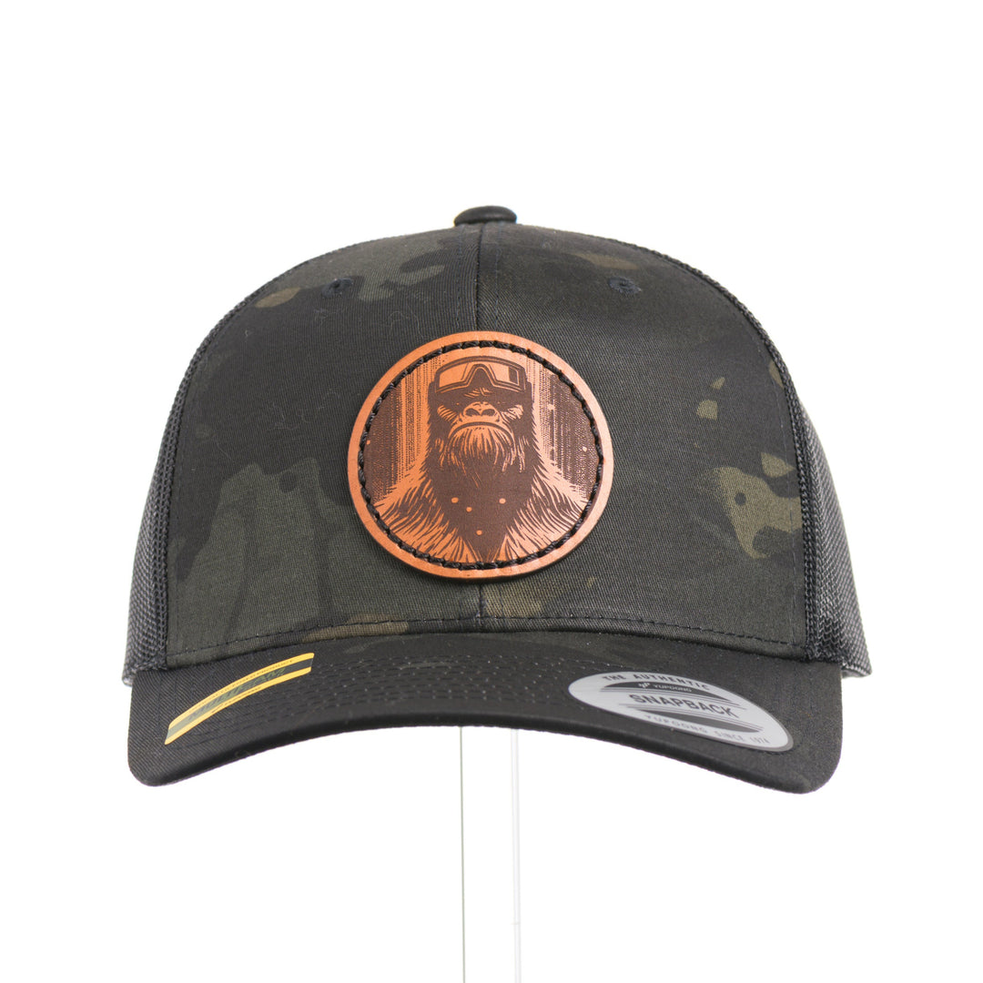 The front of a Snowy Bigfoot Leather Patch Black Camo Trucker Hat, featuring a circular real hand-dyed leather patch etched with a detailed Bigfoot in ski googles design, securely heat bonded and sewn onto the structured front of a dark camo Yupoong Retro Trucker Hat—handmade in Montana, USA by Last Best Supply Co.