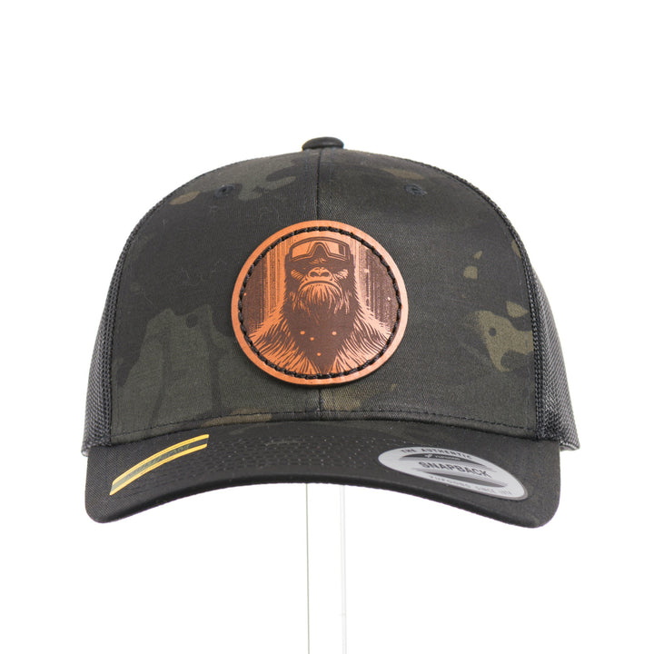 The front of a Snowy Bigfoot Leather Patch Black Camo Trucker Hat, featuring a circular real hand-dyed leather patch etched with a detailed Bigfoot in ski googles design, securely heat bonded and sewn onto the structured front of a dark camo Yupoong Retro Trucker Hat—handmade in Montana, USA by Last Best Supply Co.