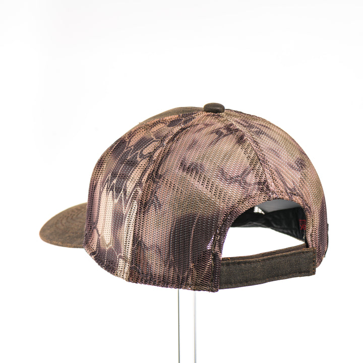 The back of a Walnut & Bamboo Copper Trout Patch Kryptek Camo Trucker Hat, featuring a rectangular real walnut wood veneer patch with cut-out trout silhouette and bamboo-colored copper inlay, laminated to a black anodized metal plate, and riveted to a chocolate brown and Kryptek camo mesh trucker hat—handmade in Montana, USA by Last Best Supply Co