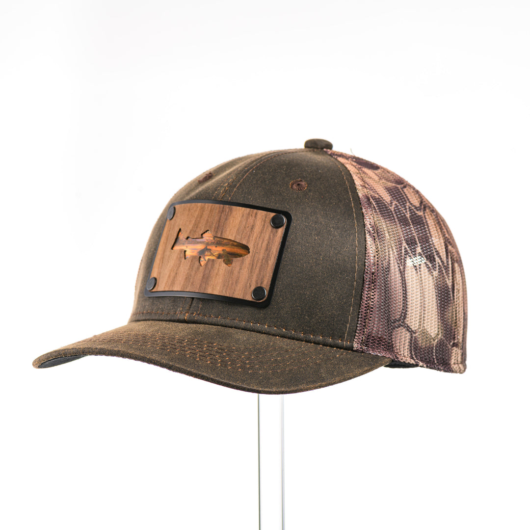 3/4 view of a Walnut & Bamboo Copper Trout Patch Kryptek Camo Trucker Hat, featuring a rectangular real walnut wood veneer patch with cut-out trout silhouette and bamboo-colored copper inlay, laminated to a black anodized metal plate, and riveted to a chocolate brown and Kryptek camo mesh trucker hat—handmade in Montana, USA by Last Best Supply Co.