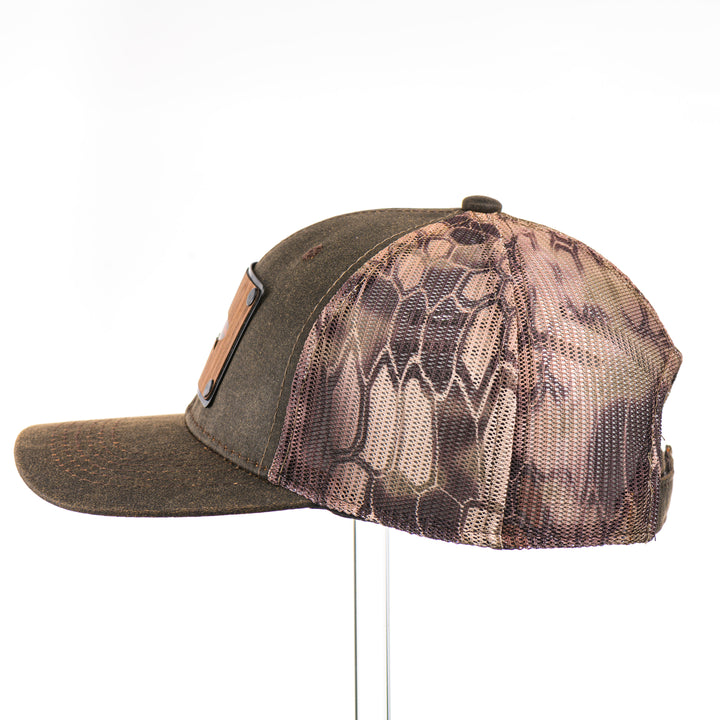 The side of a Walnut & Bamboo Copper Trout Patch Kryptek Camo Trucker Hat, featuring a rectangular real walnut wood veneer patch with cut-out trout silhouette and bamboo-colored copper inlay, laminated to a black anodized metal plate, and riveted to a chocolate brown and Kryptek camo mesh trucker hat—handmade in Montana, USA by Last Best Supply Co.