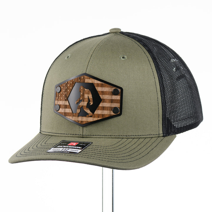 Walnut Wood Bigfoot American Flag Patch Trucker Hat featuring a real walnut wood patch laminated to a hexagon-shaped black anodized aluminum plate with an etched American flag and Bigfoot design, handmade in Montana, USA by Last Best Supply Co.