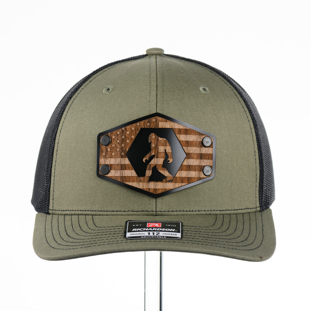 Walnut Wood Bigfoot American Flag Patch Trucker Hat featuring a real walnut wood patch laminated to a hexagon-shaped black anodized aluminum plate with an etched American flag and Bigfoot design, handmade in Montana, USA by Last Best Supply Co.