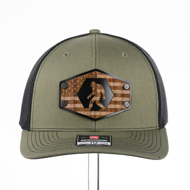 Walnut Wood Bigfoot American Flag Patch Trucker Hat featuring a real walnut wood patch laminated to a hexagon-shaped black anodized aluminum plate with an etched American flag and Bigfoot design, handmade in Montana, USA by Last Best Supply Co.
