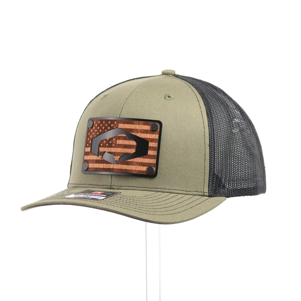 A loden green and black mesh Richardson trucker hat with an etched walnut wood American flag and bison cut out on a black metal patch at a quarter turn—handmade in Montana, USA by Last Best Supply Co.