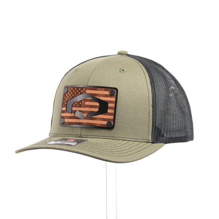 A loden green and black mesh Richardson trucker hat with an etched walnut wood American flag and bison cut out on a black metal patch at a quarter turn—handmade in Montana, USA by Last Best Supply Co.