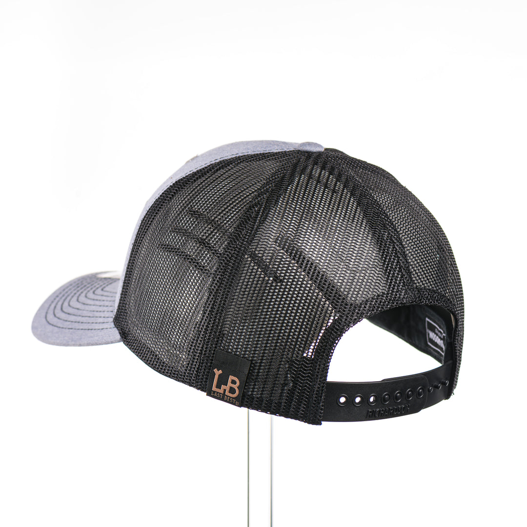 The back of a heather grey and black mesh Richardson trucker hat featuring a real walnut wood patch laser-etched with the American flag with a cut-out Chinook salmon silhouette, layered over a black hexagon-shaped anodized aluminum plate—handmade in Montana, USA by Last Best Supply Co.