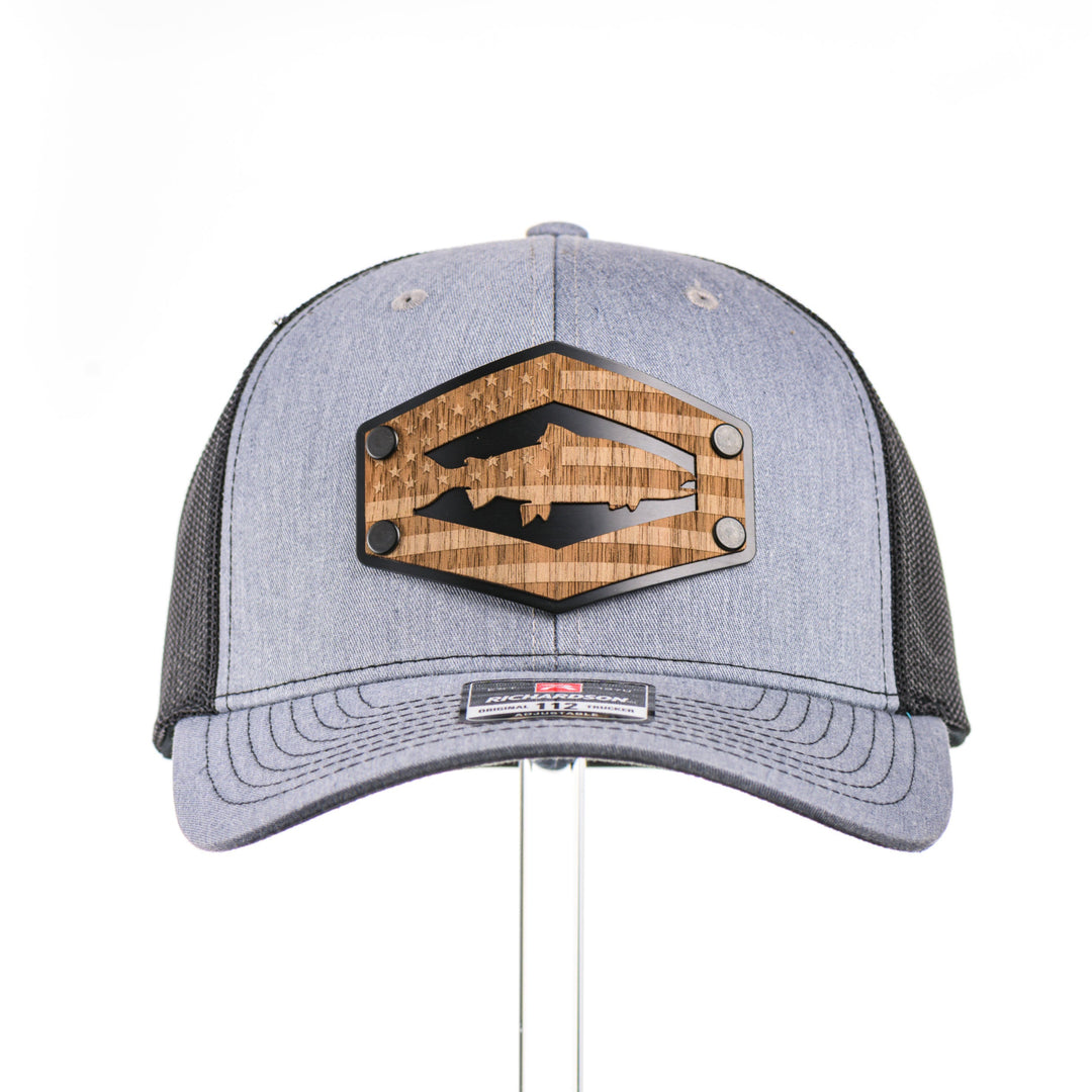The front of a heather grey and black mesh Richardson trucker hat featuring a real walnut wood patch laser-etched with the American flag with a cut-out Chinook salmon silhouette, layered over a black hexagon-shaped anodized aluminum plate—handmade in Montana, USA by Last Best Supply Co.