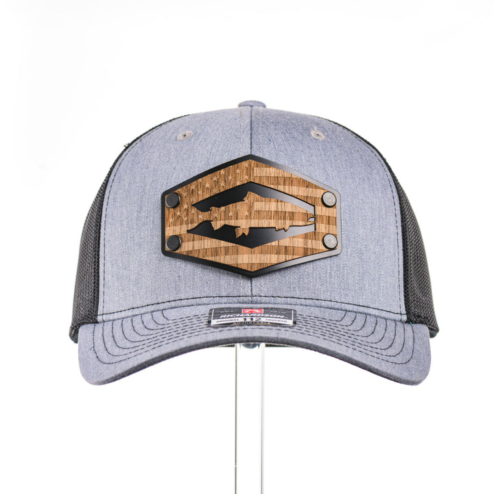 The front of a heather grey and black mesh Richardson trucker hat featuring a real walnut wood patch laser-etched with the American flag with a cut-out Chinook salmon silhouette, layered over a black hexagon-shaped anodized aluminum plate—handmade in Montana, USA by Last Best Supply Co.