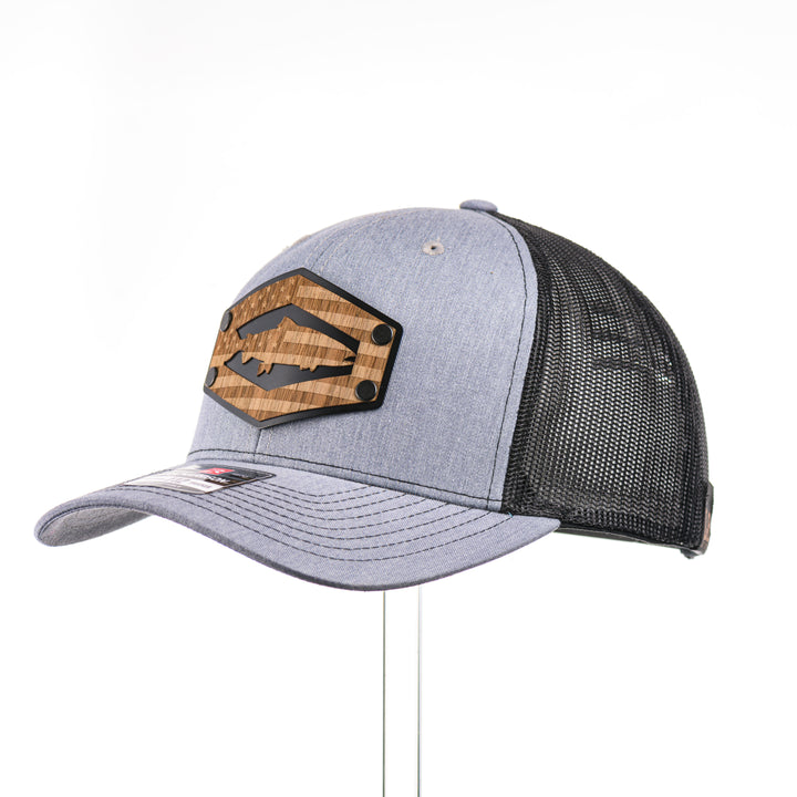 3/4 view of a heather grey and black mesh Richardson trucker hat featuring a real walnut wood patch laser-etched with the American flag with a cut-out Chinook salmon silhouette, layered over a black hexagon-shaped anodized aluminum plate—handmade in Montana, USA by Last Best Supply Co.