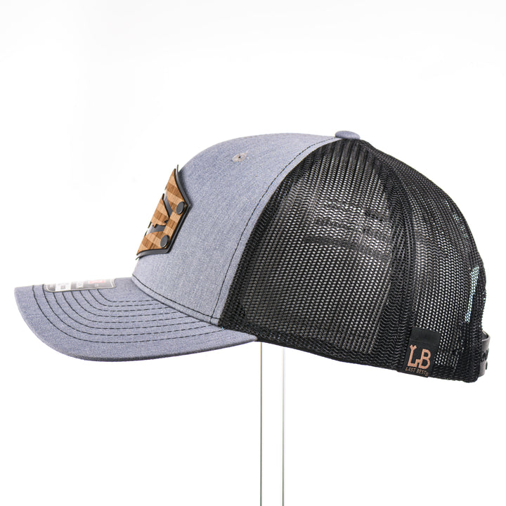 The side view of a heather grey and black mesh Richardson trucker hat featuring a real walnut wood patch laser-etched with the American flag with a cut-out Chinook salmon silhouette, layered over a black hexagon-shaped anodized aluminum plate—handmade in Montana, USA by Last Best Supply Co.
