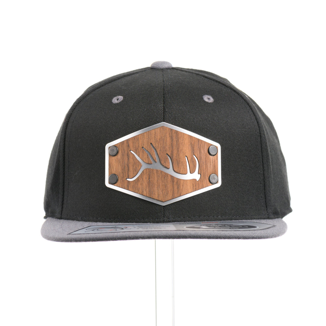 The front of a Walnut Wood Tines Flexfit Black & Grey Flat Bill Hat, featuring a real walnut wood veneer patch precision-cut into a hexagon with bold cut-out elk antler tines, laminated to a hexagon silver anodized aluminum plate, and riveted to a heather gray and black Flexfit 110F trucker hat—handmade in Montana, USA by Last Best Supply Co.