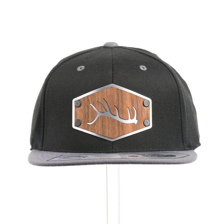 The front of a Walnut Wood Tines Flexfit Black & Grey Flat Bill Hat, featuring a real walnut wood veneer patch precision-cut into a hexagon with bold cut-out elk antler tines, laminated to a hexagon silver anodized aluminum plate, and riveted to a heather gray and black Flexfit 110F trucker hat—handmade in Montana, USA by Last Best Supply Co.
