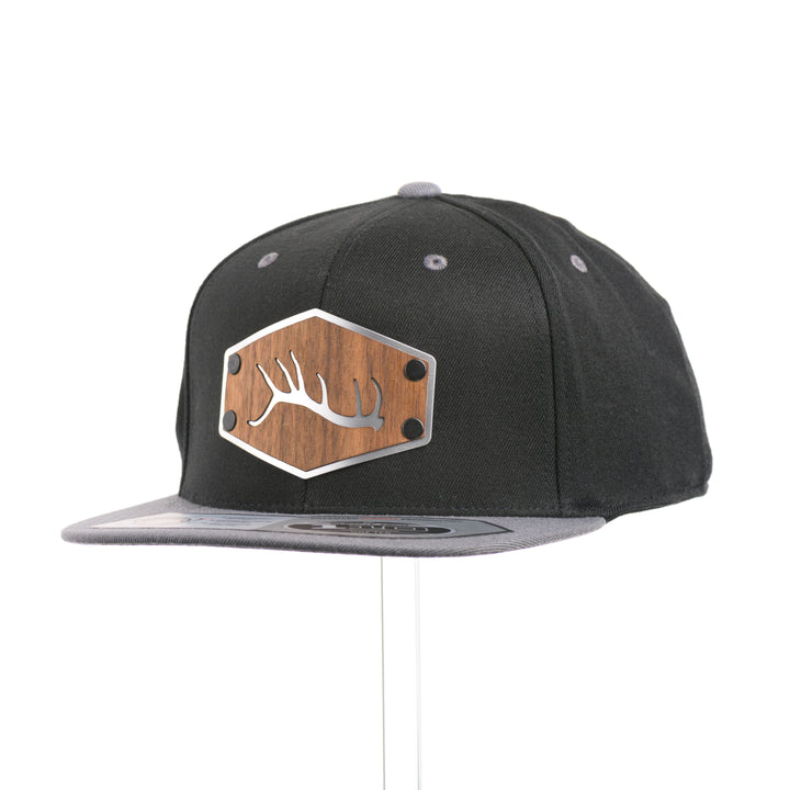 3/4 view of a Walnut Wood Tines Flexfit Black & Grey Flat Bill Hat, featuring a real walnut wood veneer patch precision-cut into a hexagon with bold cut-out elk antler tines, laminated to a hexagon silver anodized aluminum plate, and riveted to a heather gray and black Flexfit 110F trucker hat—handmade in Montana, USA by Last Best Supply Co.