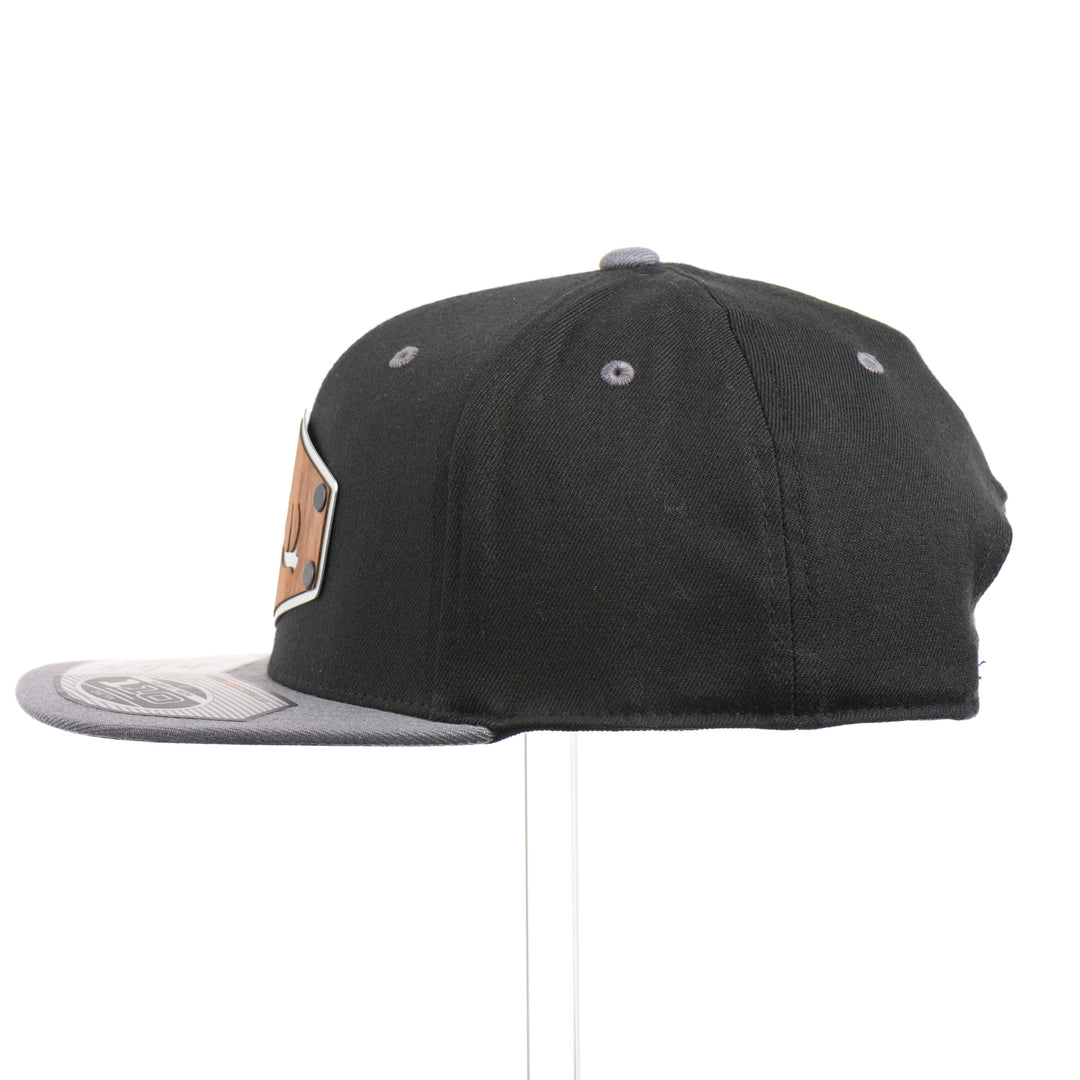 The side of a Walnut Wood Tines Flexfit Black & Grey Flat Bill Hat, featuring a real walnut wood veneer patch precision-cut into a hexagon with bold cut-out elk antler tines, laminated to a hexagon silver anodized aluminum plate, and riveted to a heather gray and black Flexfit 110F trucker hat—handmade in Montana, USA by Last Best Supply Co.