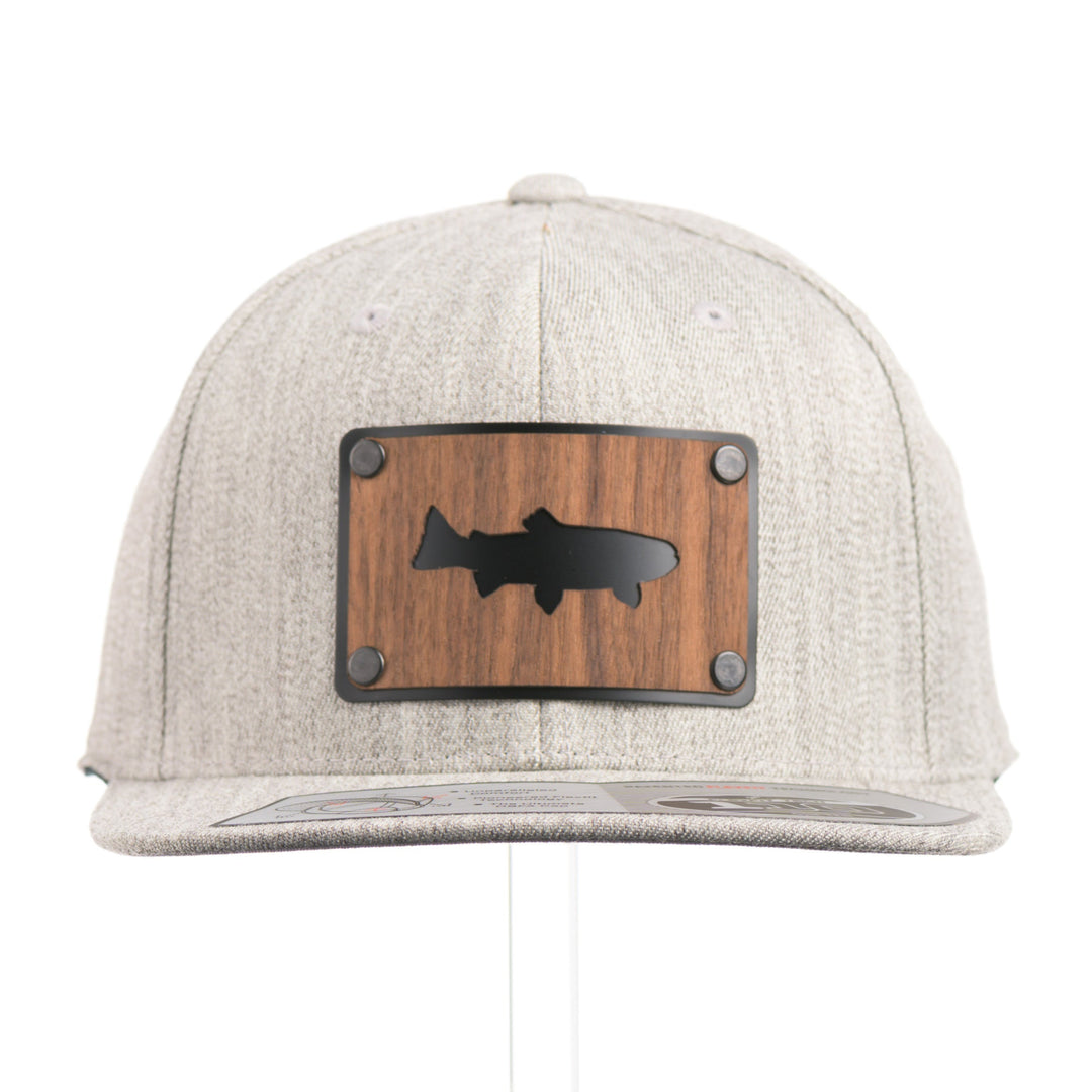 The front of a Walnut Trout Patch Plate Heather Grey Flat Bill Hat, featuring a rectangular real walnut wood veneer patch with cut-out trout silhouette, laminated to a black anodized metal plate, and riveted to a light heather grey 6-panel Flexfit Flat Bill Hat—handmade in Montana, USA by Last Best Supply Co.