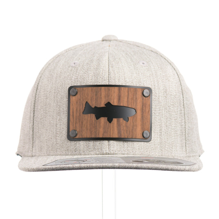 The front of a Walnut Trout Patch Plate Heather Grey Flat Bill Hat, featuring a rectangular real walnut wood veneer patch with cut-out trout silhouette, laminated to a black anodized metal plate, and riveted to a light heather grey 6-panel Flexfit Flat Bill Hat—handmade in Montana, USA by Last Best Supply Co.