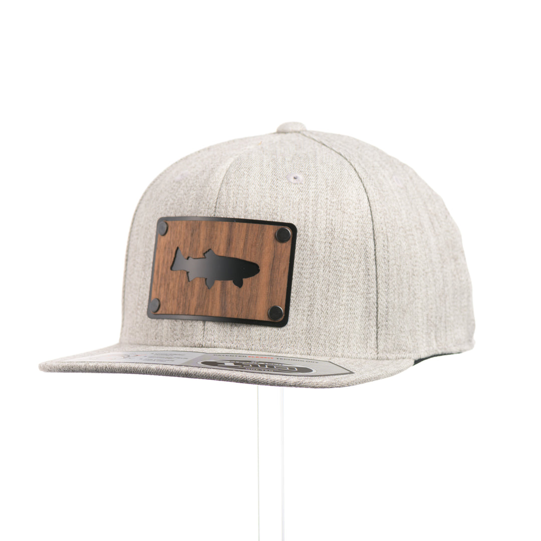 3/4 view of a Walnut Trout Patch Plate Heather Grey Flat Bill Hat, featuring a rectangular real walnut wood veneer patch with cut-out trout silhouette, laminated to a black anodized metal plate, and riveted to a light heather grey 6-panel Flexfit Flat Bill Hat—handmade in Montana, USA by Last Best Supply Co.