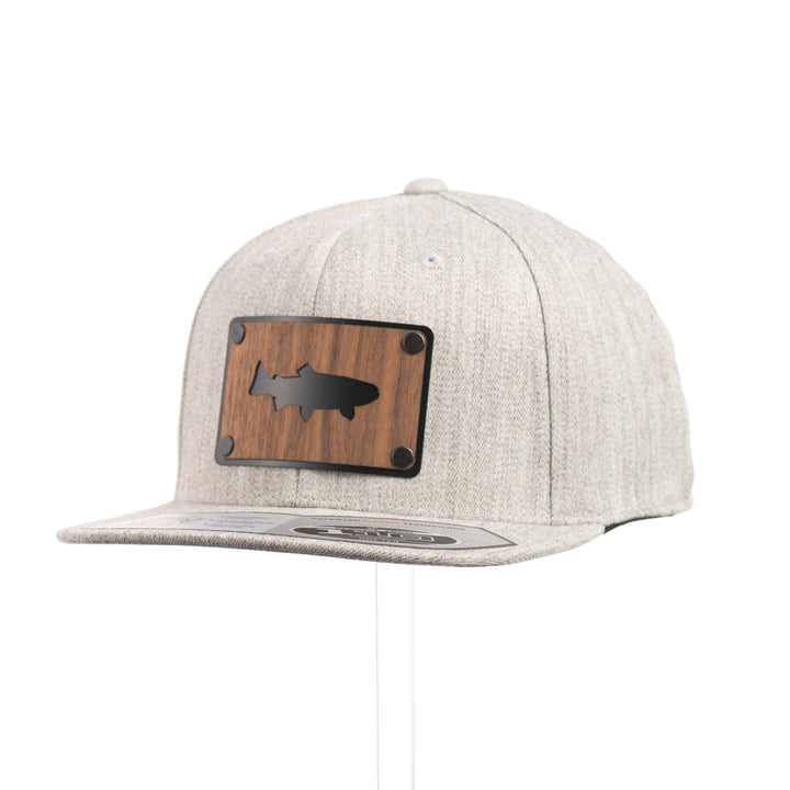 3/4 view of a Walnut Trout Patch Plate Heather Grey Flat Bill Hat, featuring a rectangular real walnut wood veneer patch with cut-out trout silhouette, laminated to a black anodized metal plate, and riveted to a light heather grey 6-panel Flexfit Flat Bill Hat—handmade in Montana, USA by Last Best Supply Co.