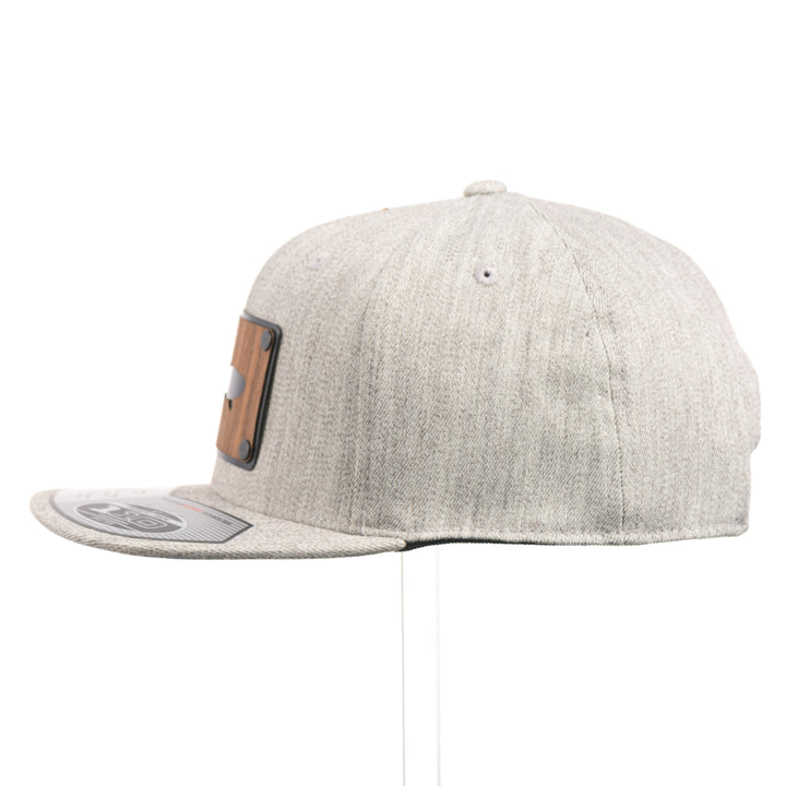 The side of a Walnut Trout Patch Plate Heather Grey Flat Bill Hat, featuring a rectangular real walnut wood veneer patch with cut-out trout silhouette, laminated to a black anodized metal plate, and riveted to a light heather grey 6-panel Flexfit Flat Bill Hat—handmade in Montana, USA by Last Best Supply Co.