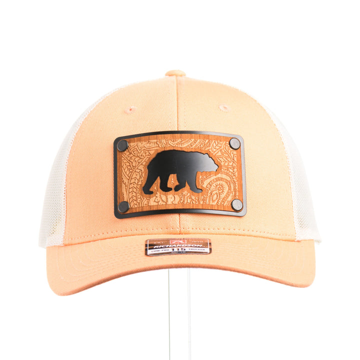 The front of a Cherry Wood Grizzly Bear w/ Paisley Peach & Cream Trucker Hat, featuring a real cherry wood veneer patch etched with a paisley design around a bear silhouette cut-out, laminated to a black anodized aluminum plate, and riveted to a peaches-and-cream Richardson 115 low-profile trucker hat—handmade in Montana, USA by Last Best Supply Co.