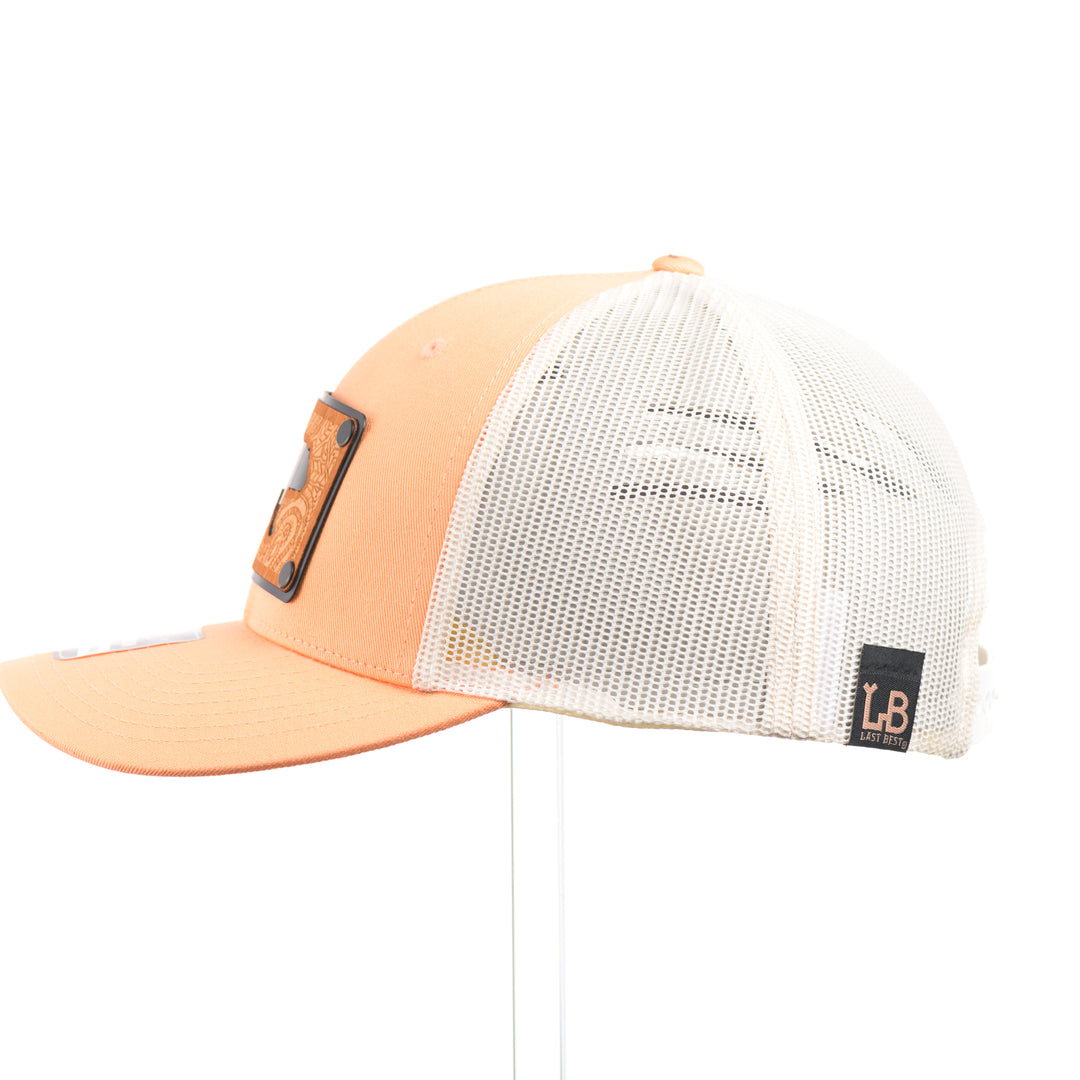 The side of a Cherry Wood Grizzly Bear w/ Paisley Peach & Cream Trucker Hat, featuring a real cherry wood veneer patch etched with a paisley design around a bear silhouette cut-out, laminated to a black anodized aluminum plate, and riveted to a peaches-and-cream Richardson 115 low-profile trucker hat—handmade in Montana, USA by Last Best Supply Co.